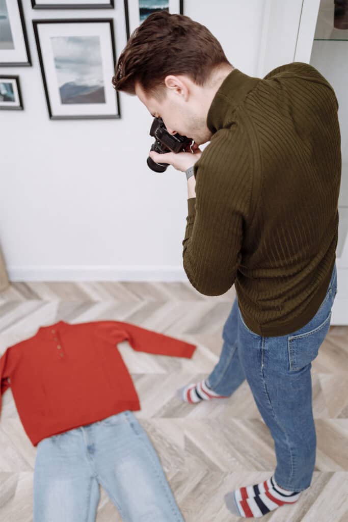 man in turtleneck and jeans taking a picture of a sweater and jeans laying on the floor