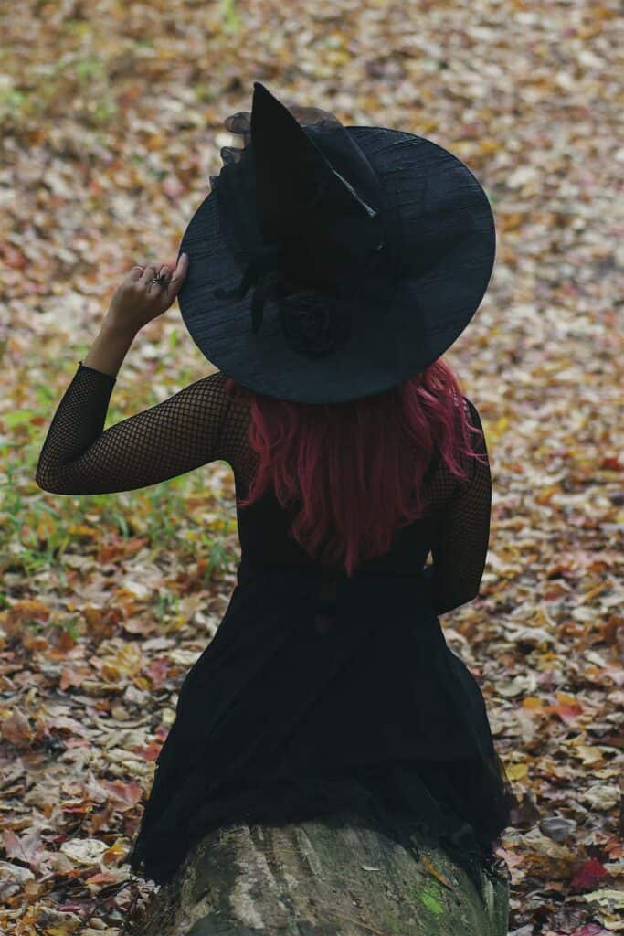 woman in an adult halloween costume with a witch hat sitting in a pile of leaves.