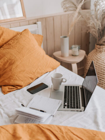 a laptop, notebook, phone, and coffee mug sitting on a bed with white sheets and orange pillows.