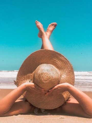 person in a tan straw floppy hat laying on the beach with the ocean in front.