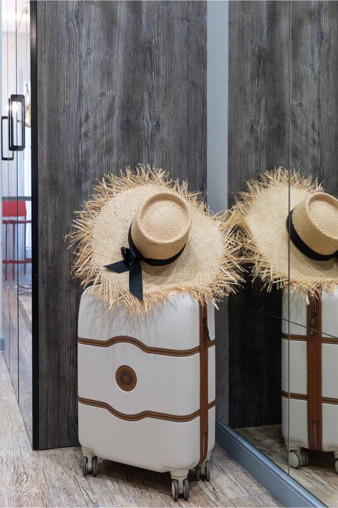 a white suitcase with 4 wheels with brown detailing with a tan straw hat sitting on top of the suitcase.