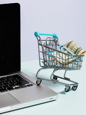 a small shopping cart with folded money inside of it next to a black and silver laptop.