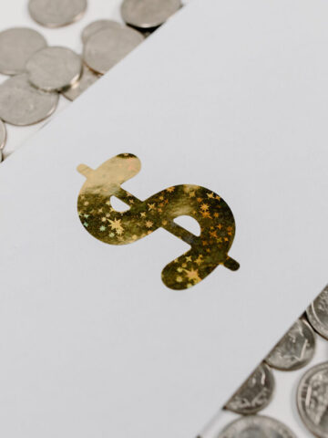 a gold sparkly dollar sign on a white piece of paper laying ontop of quarters scattered across a table.