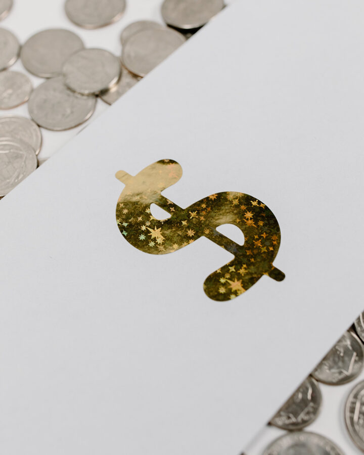 a gold sparkly dollar sign on a white piece of paper laying ontop of quarters scattered across a table.