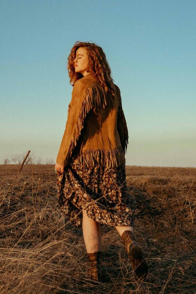 back view of a girl standing in a field wearing a suede fringe jacket.