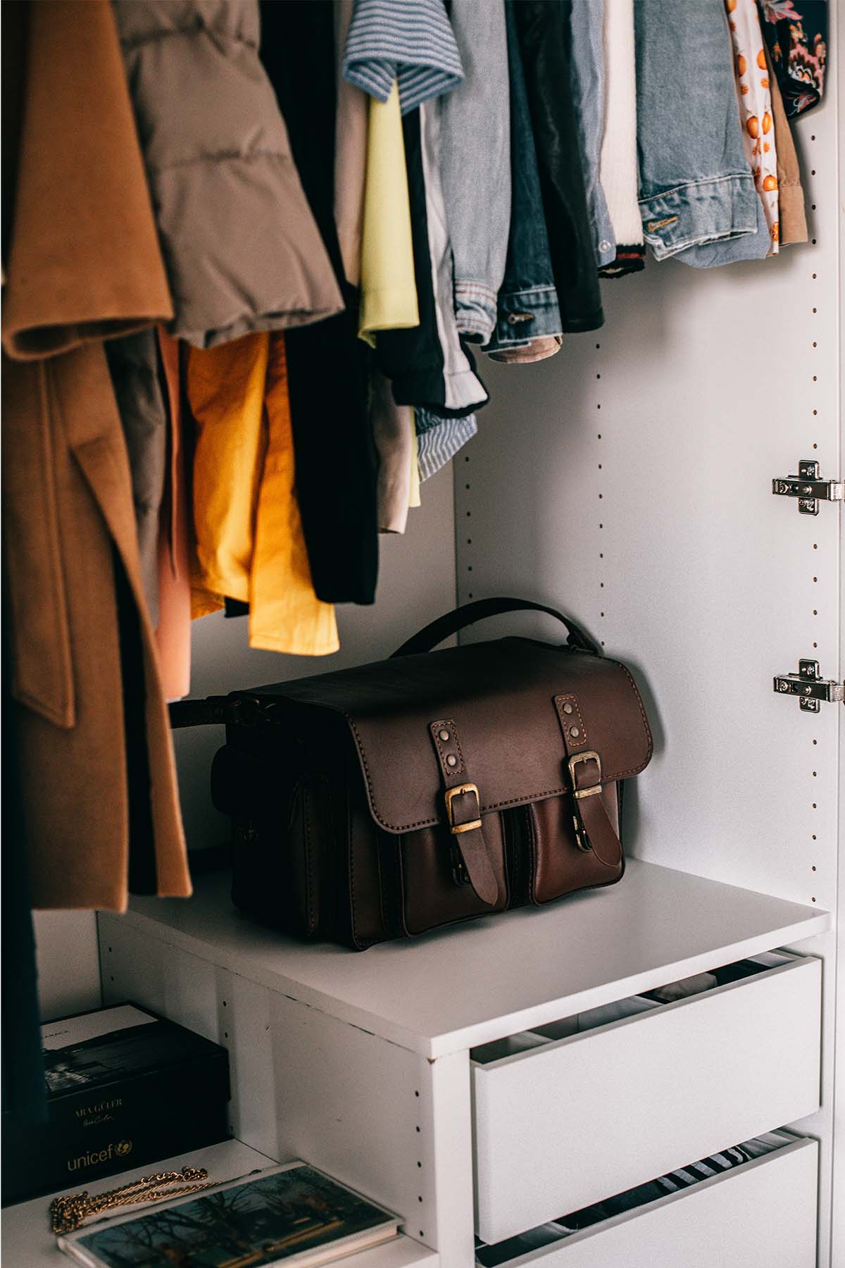 a closet with clothes hanging and a brown purse sitting on a shelf.